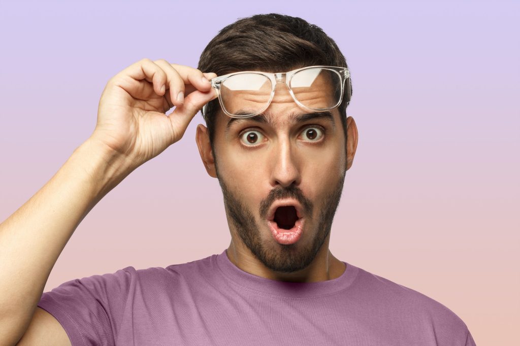 Young man in t-shirt shouting OMG with open mouth, surprised by sales, holding transparent glasses