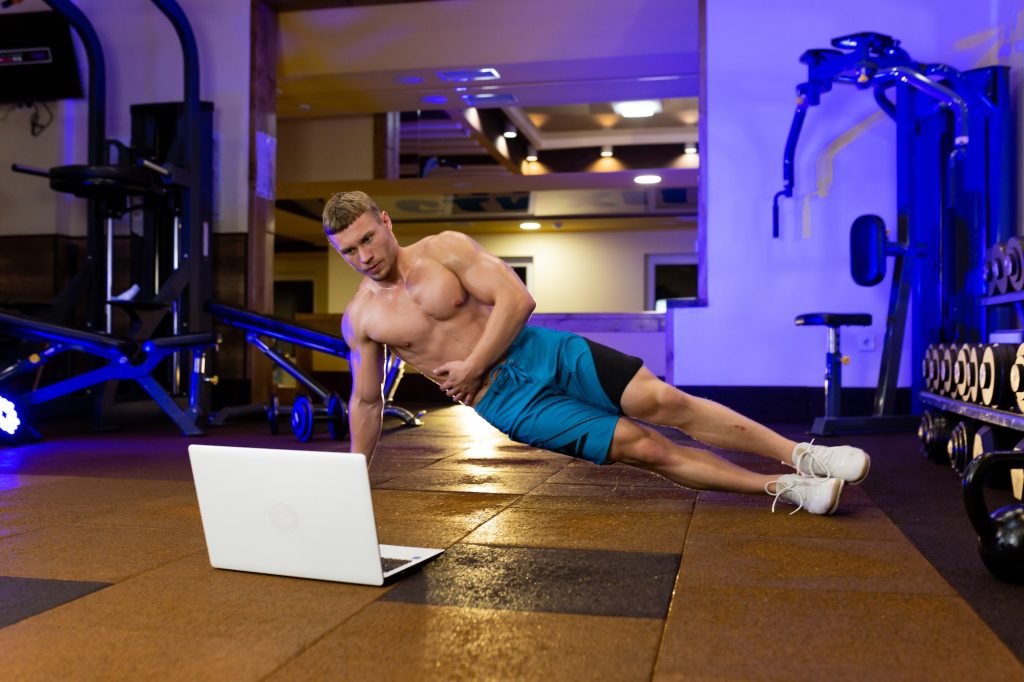 Young sports fitness trainer man conducts sports training online from laptop in gym, teaches, shows